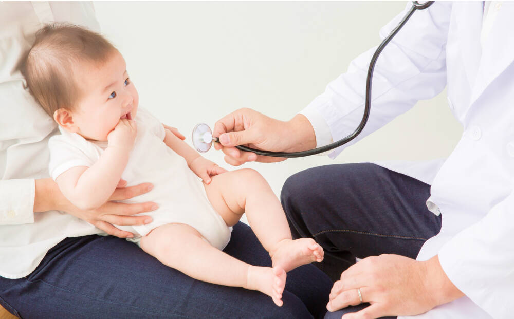 小さな赤ちゃんが保護者の膝の上に座り、医師が聴診器で診察をしている様子